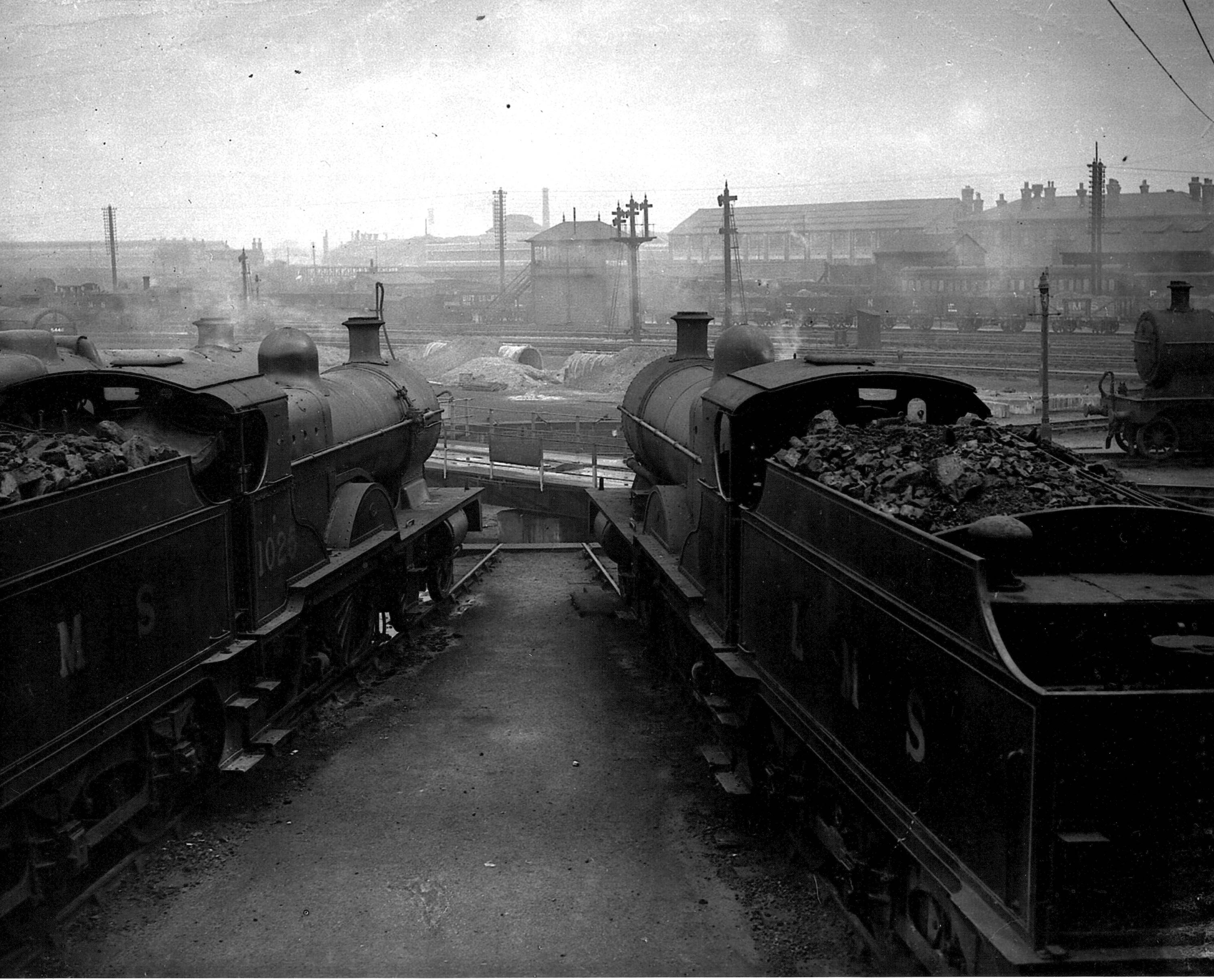 A photograph taken with two locomotives in the foreground stabled around a turntable at Derby's 'Four Shed'. We are looking over to Way & Works signal box with air raid shelters being contructed in the middle-distance. The LMS research laboratories on London Road are visible in the background.