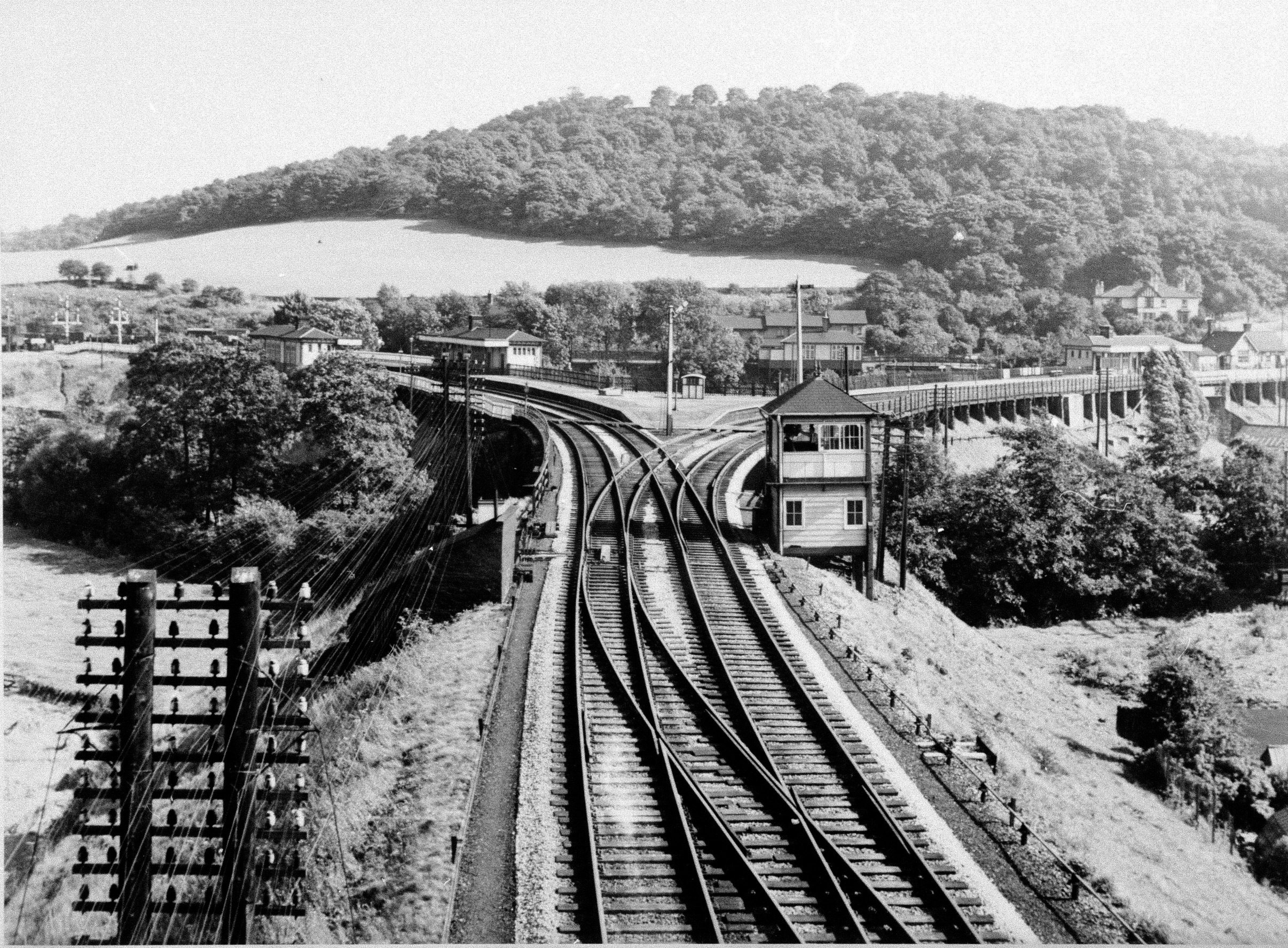 Two railway tracks curve through a lush landscape with a tunnel, signal boxes, and a distant body of water under a clear sky.