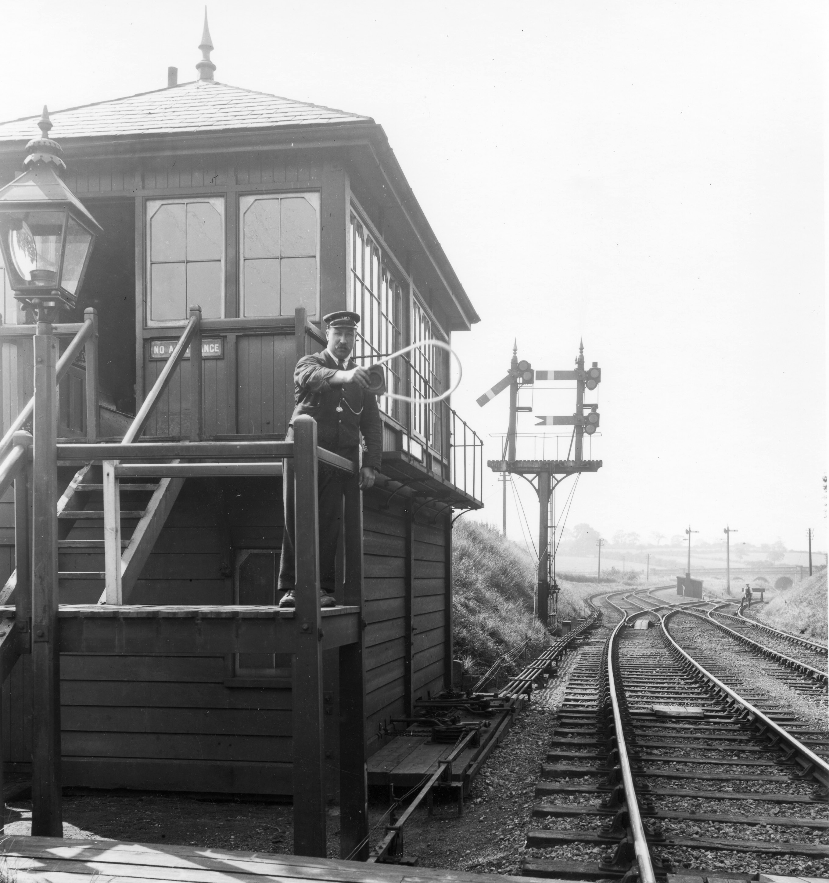 A LMS signalman posing on the steps of his signal box holdong out a single line toekn for a (non-existent) train to collect.