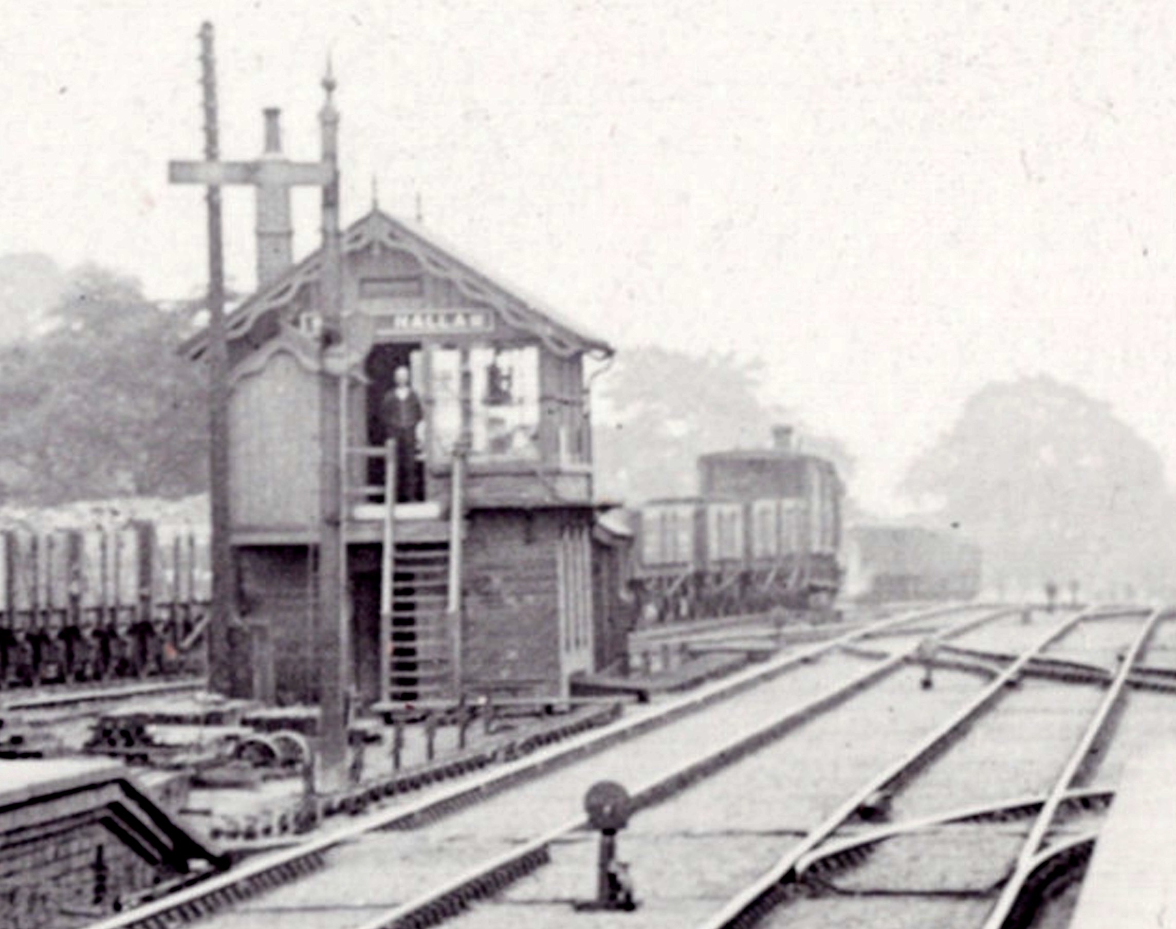 A crop from a larger image compressing the perspective, showing West Hallan GNR station signal box with the signalman standing in the doorway watching the distant photographer. The colliery sidings behind hold a rake of wagons	